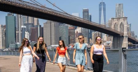 Un gruppo di cinque ragazze alla pari che camminano una accanto all'altra a Manhattan con il ponte di Brooklyn sullo sfondo.