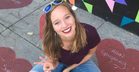 La ragazza alla pari è seduta per terra, fa il segno della pace e sorride alla telecamera.