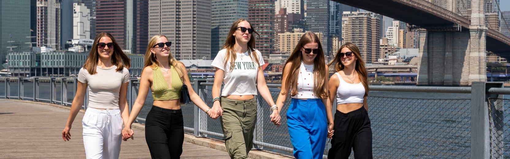 Cinque ragazze alla pari che camminano una accanto all'altra a New York con il ponte di Brooklyn sullo sfondo