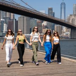 Cinque ragazze alla pari che camminano una accanto all'altra a New York con il ponte di Brooklyn sullo sfondo